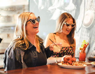 Two friends wearing heart-shaped sunglasses laugh at a bar, enjoying cocktails and a plate of fried chicken on waffles.