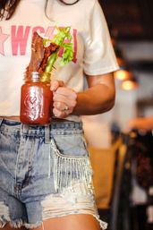 Trendy brunch scene: mason-jar Bloody Mary topped with celery stalk and crispy bacon, held by someone in distressed denim shorts and a cropped tee at a cozy café.