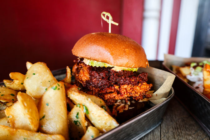 Close-up of a crispy fried chicken sandwich with pickles on a glossy brioche bun, served with seasoned potato wedges in a metal tray at a casual restaurant.