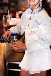 Bride-to-be in a light denim jacket and white mini skirt holding a champagne flute and a sparkly "Wifey" clutch at a bachelorette-style celebration