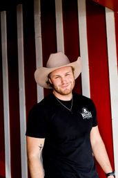 Smiling person in a beige cowboy hat and black T‑shirt standing against red-and-white wooden barn siding, casual country-style portrait with a small airplane tattoo on the forearm.