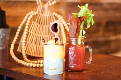 Rustic brunch scene: bloody mary in a mason jar topped with celery and crispy bacon beside a lemon cocktail in a retro tin, with woven bag and sunglasses in the background