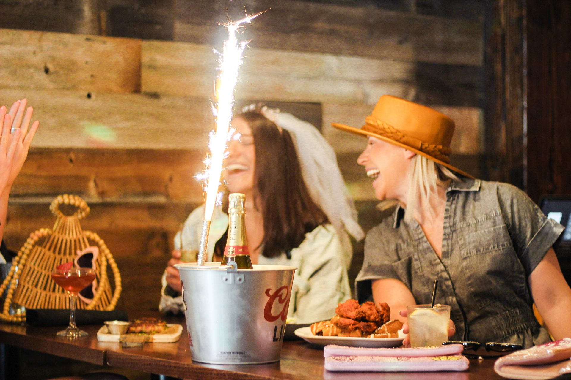Bachelorette party friends laughing at a rustic wooden bar, champagne bottle in an ice bucket with a tall sparkler, cocktails and a plate of fried chicken on the table.