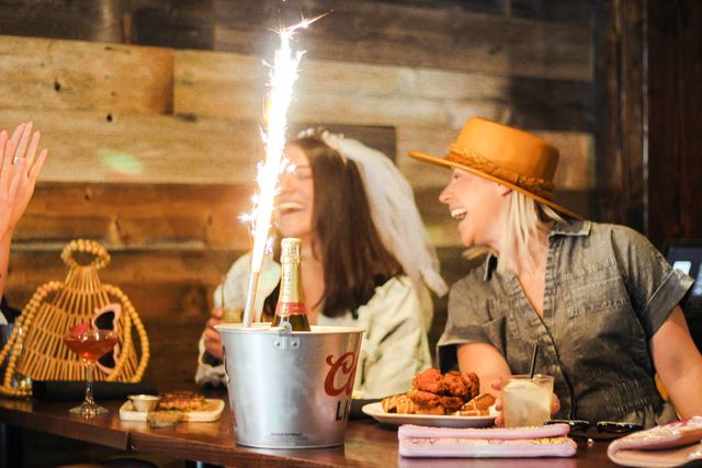 Bachelorette party friends laughing at a rustic wooden bar, champagne bottle in an ice bucket with a tall sparkler, cocktails and a plate of fried chicken on the table.