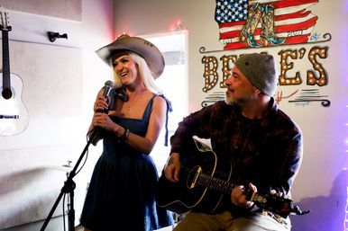 Smiling woman in a cowboy hat sings into a microphone as a bearded man in a beanie plays acoustic guitar in a cozy Americana music room with hanging guitars and a flag mural.