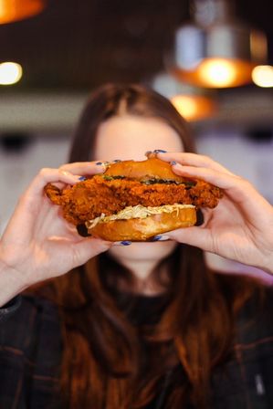 Close-up of a person holding a large crispy fried chicken sandwich with coleslaw on a brioche bun in a casual indoor restaurant, warm bokeh lights in the background