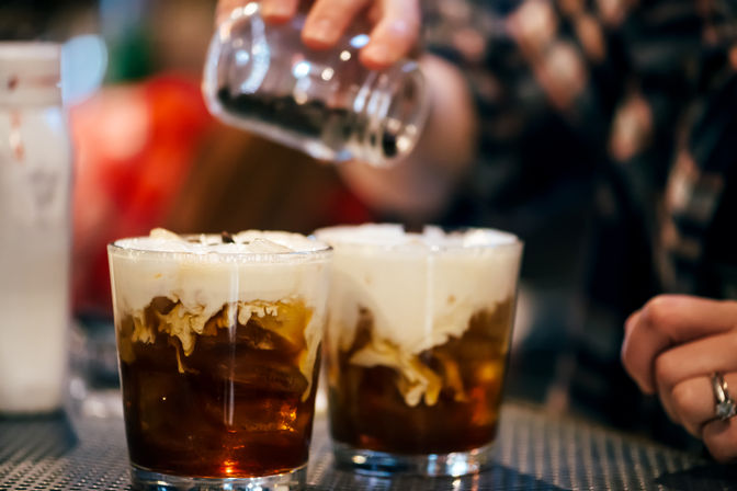 Two iced cold-brew glasses with creamy white swirls as someone sprinkles topping from a jar on a cafe counter