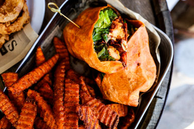Crispy seasoned crinkle-cut sweet potato fries next to an orange toasted tortilla wrap stuffed with grilled chicken and fresh romaine, secured with a wooden pick in a metal serving tray.