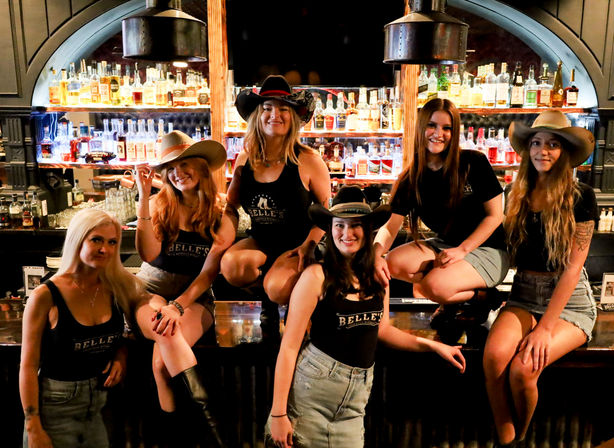 Six smiling women in cowgirl hats and denim posing on a rustic saloon-style bar counter with backlit liquor bottles