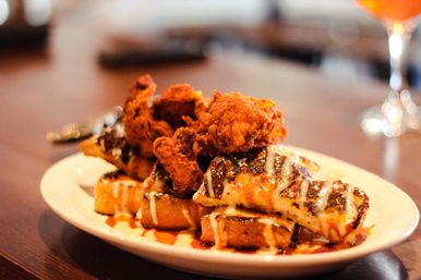 Crispy fried chicken perched on golden grilled waffles, drizzled with syrup and creamy glaze on a white plate on a wooden table — classic chicken-and-waffles brunch