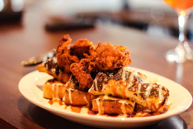 Crispy fried chicken perched on golden grilled waffles, drizzled with syrup and creamy glaze on a white plate on a wooden table — classic chicken-and-waffles brunch