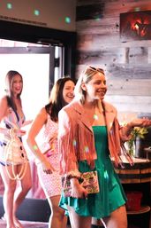 Three smiling women in colorful summer dresses, one wearing a pink fringe jacket, entering a rustic wood-paneled bar or brunch spot while holding patterned clutches and laughing under disco light reflections.