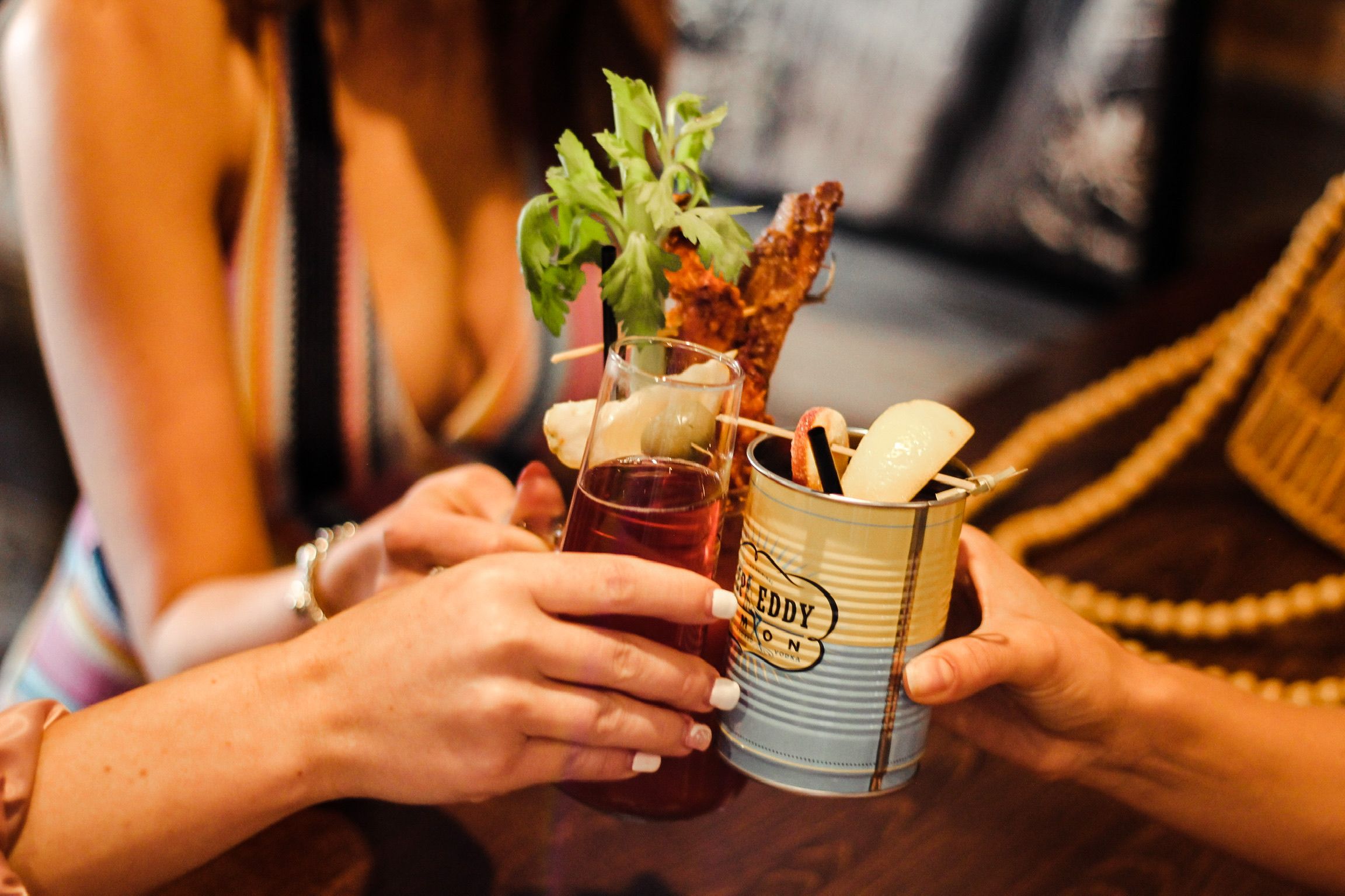 Close-up of hands clinking a tall red cocktail and a retro tin-cup drink garnished with celery, crispy bacon, pickles and a lemon wedge over a wooden bar