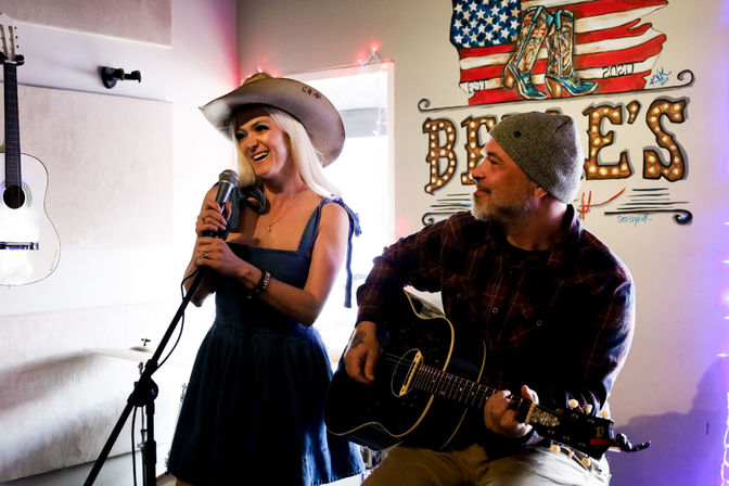 Smiling female country singer in a cowboy hat and denim dress holds a microphone beside a bearded guitarist in a beanie playing acoustic guitar in an intimate indoor country-music venue with patriotic wall art.