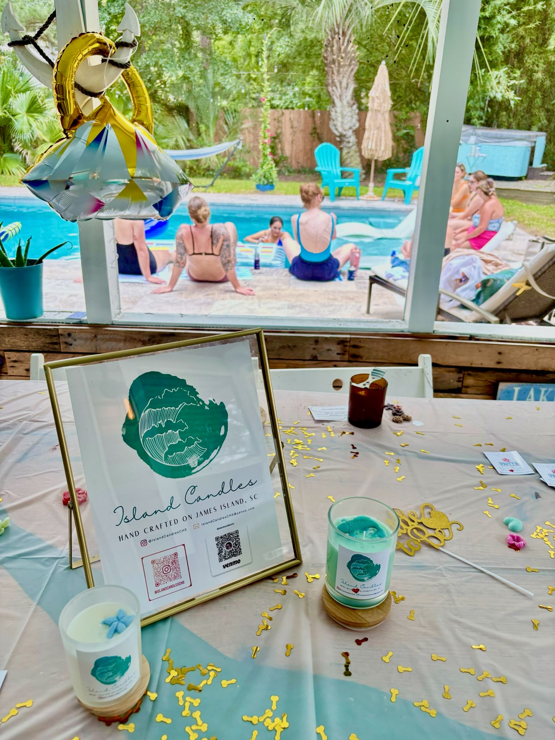 Framed candle display and teal-scented candles on a confetti-covered table at a sunny backyard pool party with friends lounging by a turquoise pool and palm trees