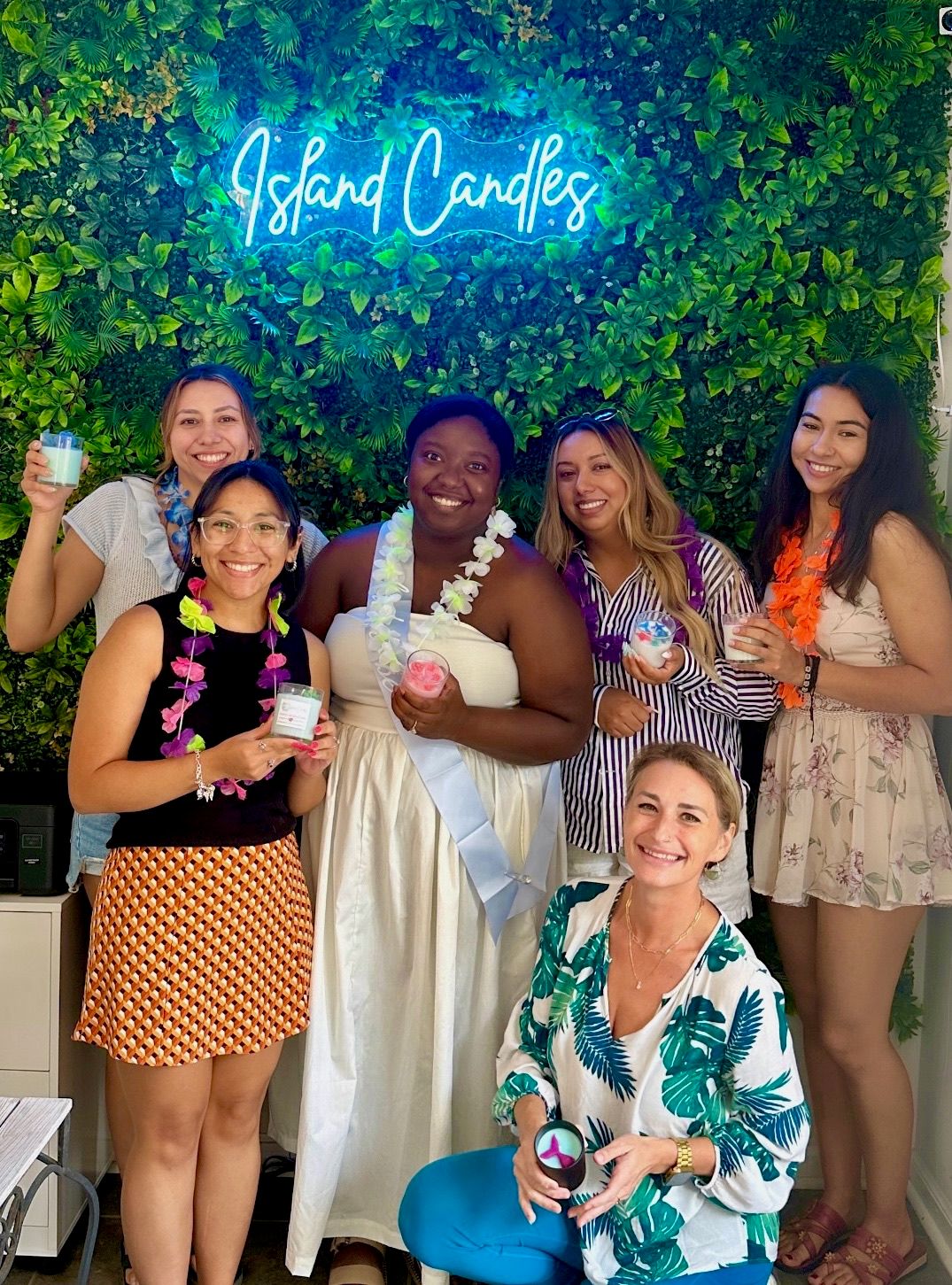 Six women smiling in a tropical-themed candle boutique under a neon cursive sign and lush green wall, wearing leis and holding colorful candles — one guest in a white dress with a sash.