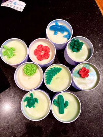 Hand-poured soy candles in white tins topped with colorful wax shapes — turtles, cactus, blue koi, red hibiscus, succulents and a fern — arranged on a dark stone countertop