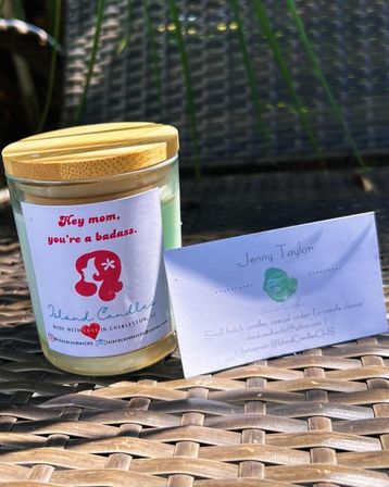 Gift candle in a glass jar with a bamboo lid labeled "Hey mom, you're a badass," sitting next to a small handwritten note on a sunlit wicker patio table.