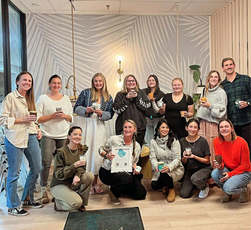 Smiling team holding coffee cups in a bright modern office lounge with a palm-leaf mural, hanging light fixture and potted plants — casual group gathering.
