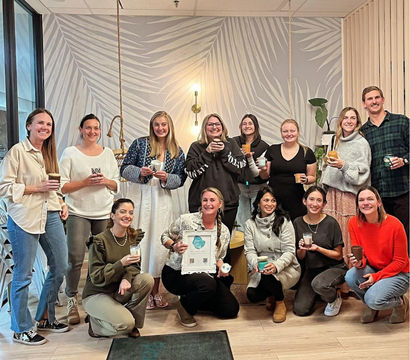 Smiling team holding coffee cups in a bright modern office lounge with a palm-leaf mural, hanging light fixture and potted plants — casual group gathering.