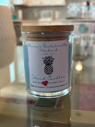 Clear glass jar candle with blue wax and bamboo lid, pineapple label on the front, sitting on a glass table in a bright kitchen — cheerful home decor or party-favor candle