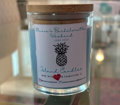 Clear glass jar candle with blue wax and bamboo lid, pineapple label on the front, sitting on a glass table in a bright kitchen — cheerful home decor or party-favor candle