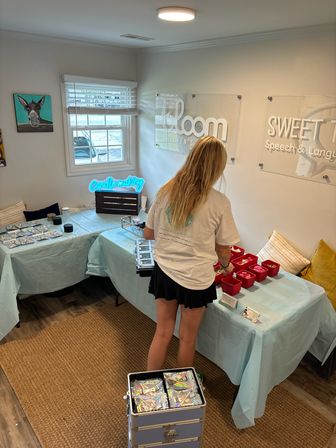 Person at a candle-making craft table in a bright small studio — teal-covered tables with red supply bins, neon candle-making sign, wall art and a daylight window.