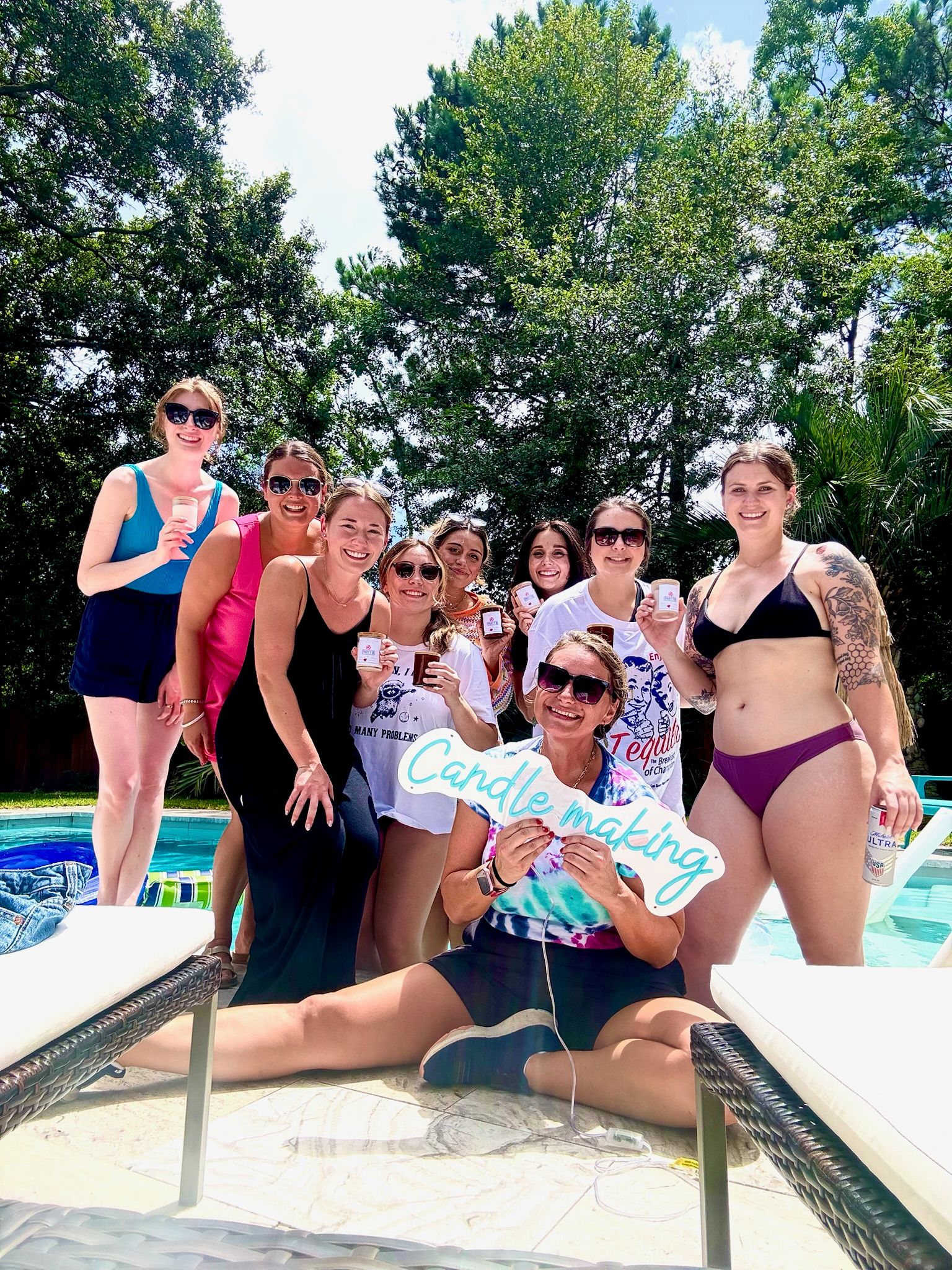 Smiling group of friends at a sunny backyard poolside candle-making party, holding mini candles and a 'Candle making' sign under tall trees.