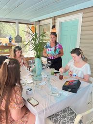 Group DIY craft workshop on a screened porch—women gathered around a plastic-covered table with craft supplies, confetti, drinks, a ceramic fish planter and turquoise door.