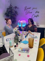 Two friends clinking tall drinks over a white table in a colorful DIY craft studio, surrounded by paints, candles, molds, tools, flowers and a neon sign.
