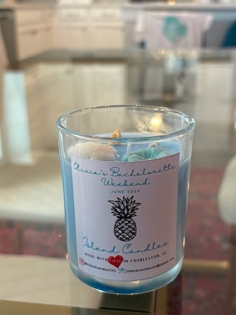Blue coastal-scented candle in a clear glass jar with a pineapple illustration and 'Bachelorette Weekend' label, topped with small seashell wax decorations, sitting on a glass table in a bright Charleston, SC kitchen