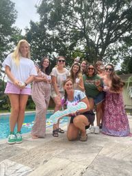 Poolside backyard candle-making party with a group of friends smiling by a pool, holding handmade candles and a neon "candle making" sign under tall trees.