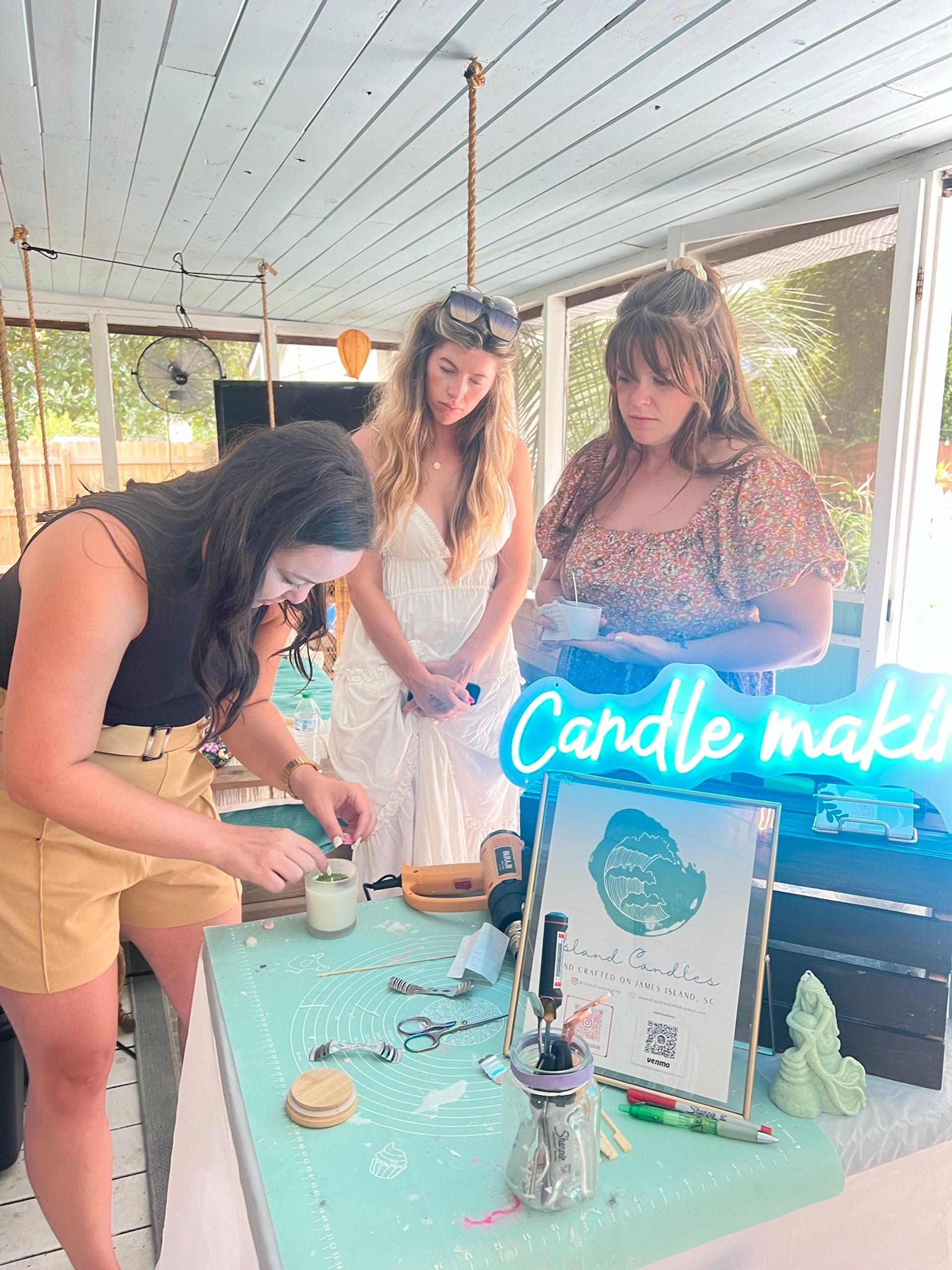 Three women at a sunny screened porch DIY candle-making workshop, one pouring wax into a jar amid tools, candles, and a glowing "Candle making" sign.