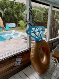 Screened tropical backyard patio overlooking a pool with turquoise chairs, in-pool loungers, palm trees, a decorative anchor, and a gold inflatable tube topped by a blue mesh pool seat.