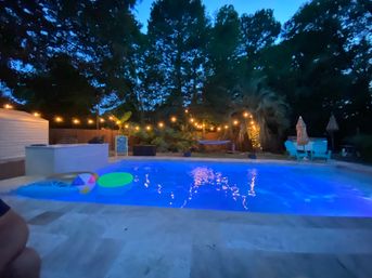 Evening backyard swimming pool with glowing blue water, string lights, inflatable floats, hammock and patio seating under palm trees.