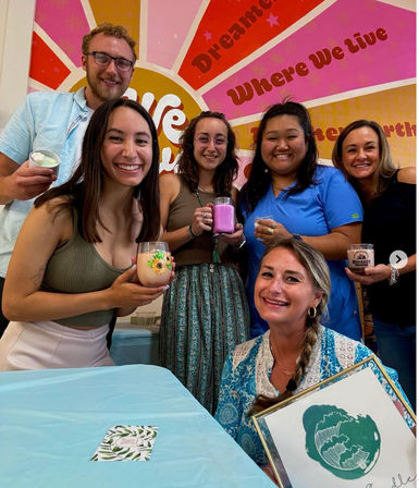 Smiling group of six adults holding handmade candles and drinks at a colorful indoor candle-making workshop in a bright craft studio with a retro sunburst mural reading ‘Where We Live’.