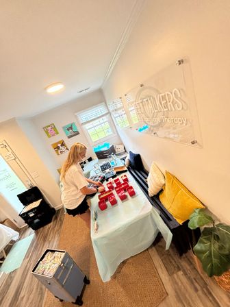 Bright, cozy speech therapy studio interior with a person arranging red activity bins on a light-blue covered table, bench with yellow pillows, colorful animal wall art, window light and a rolling storage cart.