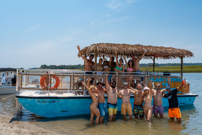 Group of people partying on a turquoise party boat with a thatched palapa roof, anchored at a shallow sandbar; everyone in swimsuits is smiling, waving and holding drinks under a clear blue sky by a marshy shoreline.