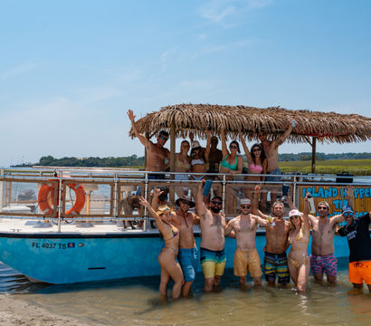 Group of people partying on a turquoise party boat with a thatched palapa roof, anchored at a shallow sandbar; everyone in swimsuits is smiling, waving and holding drinks under a clear blue sky by a marshy shoreline.