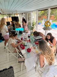 Poolside screened porch craft party: friends gathered around a white table on a covered backyard patio, working on small crafts with red containers, drinks and a potted plant centerpiece; whitewashed wood floor and ceiling, a gold ring balloon hanging and a swimming pool visible through the screens.