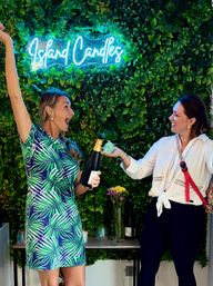 Two women celebrating in front of a lush green plant wall with a neon cursive sign; one holds a champagne bottle while the other offers a small candle, with flowers and candles on a table below.
