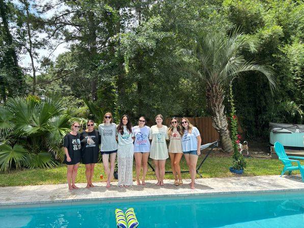 Eight women in casual summer outfits standing side-by-side by a backyard pool, surrounded by palm trees, a hammock, turquoise chairs and pool floats — relaxed summer poolside gathering.