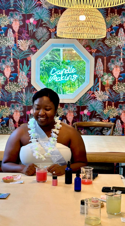 Smiling workshop participant at a candle-making class seated at a craft table with glass jars, essential oil bottles and a pink candle, colorful tropical wallpaper and a neon “Candle Making” sign in a cozy studio
