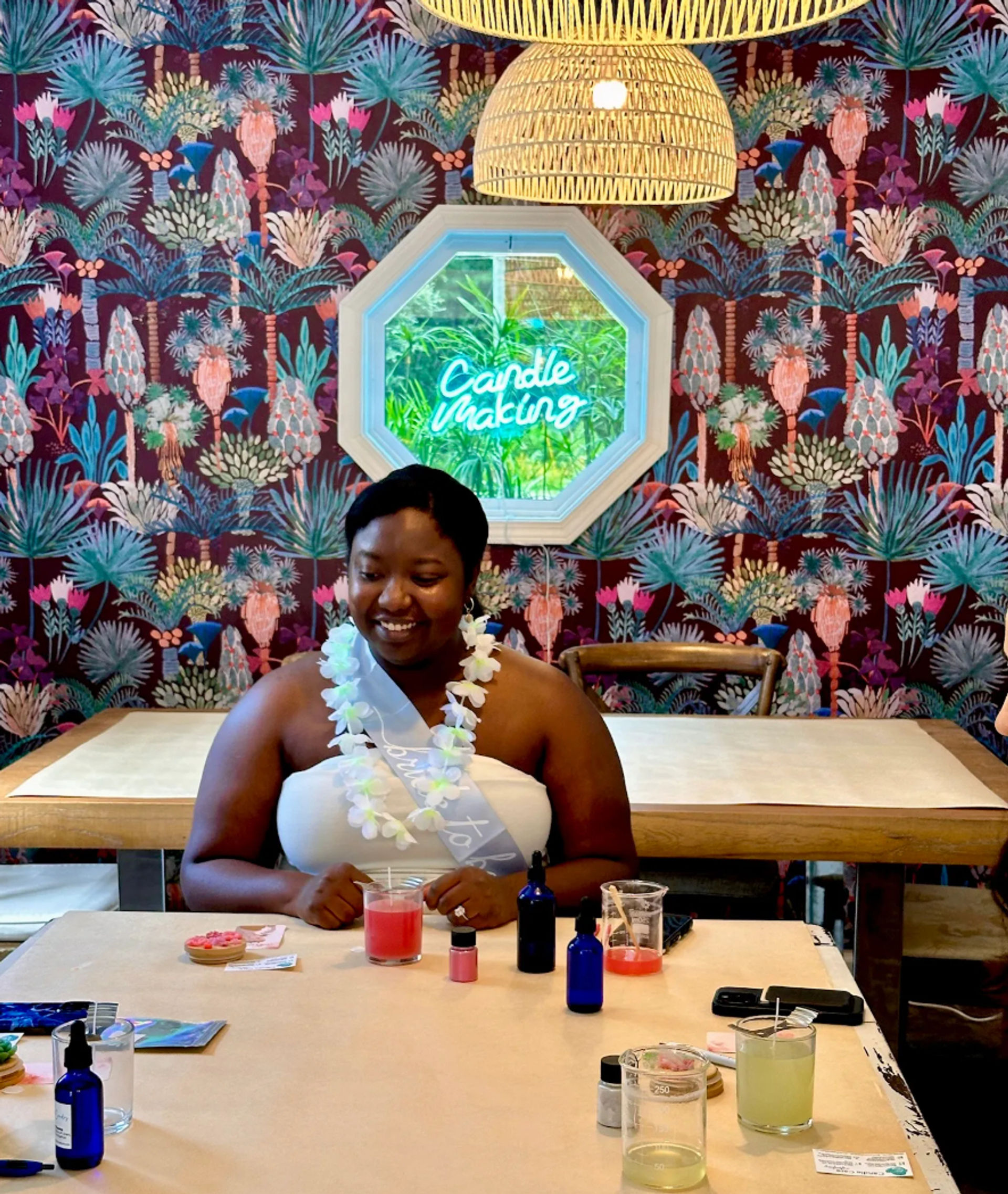 Smiling workshop participant at a candle-making class seated at a craft table with glass jars, essential oil bottles and a pink candle, colorful tropical wallpaper and a neon “Candle Making” sign in a cozy studio