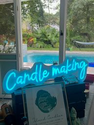 Glowing blue neon 'Candle making' sign on a table display inside a screened porch, overlooking a backyard pool, hammock and lush tropical palms.