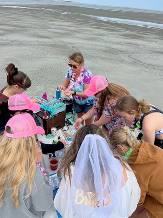 Bachelorette party on a wide sandy beach: group of women gathered around a table with craft supplies and drinks, bride wearing a white veil, several guests in pink caps and leis, ocean and low-tide flats in the background.