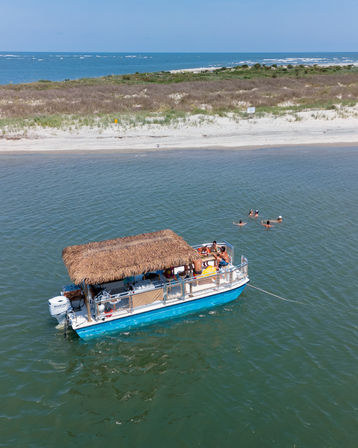 Sunny, tropical-style pontoon boat with a thatched tiki roof anchored off a sandy coastal beach, swimmers in the inlet and ocean horizon beyond.