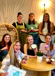 Group of smiling women at a cozy indoor candle-making party, holding handmade candles and jars around a round wooden table with a potted pink plant and rattan swing in the background.
