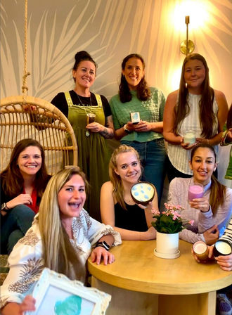 Group of smiling women at a cozy indoor candle-making party, holding handmade candles and jars around a round wooden table with a potted pink plant and rattan swing in the background.