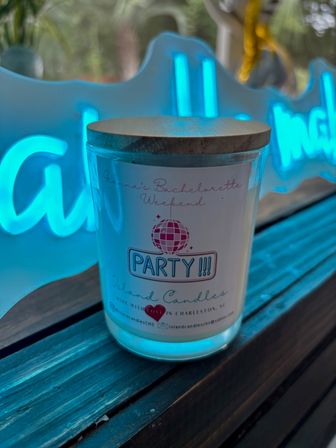 Jar candle with wooden lid labeled "PARTY!!!" and disco-ball graphic on a wooden ledge, backlit by a bright blue neon sign — party-themed candle product photo in Charleston, SC.